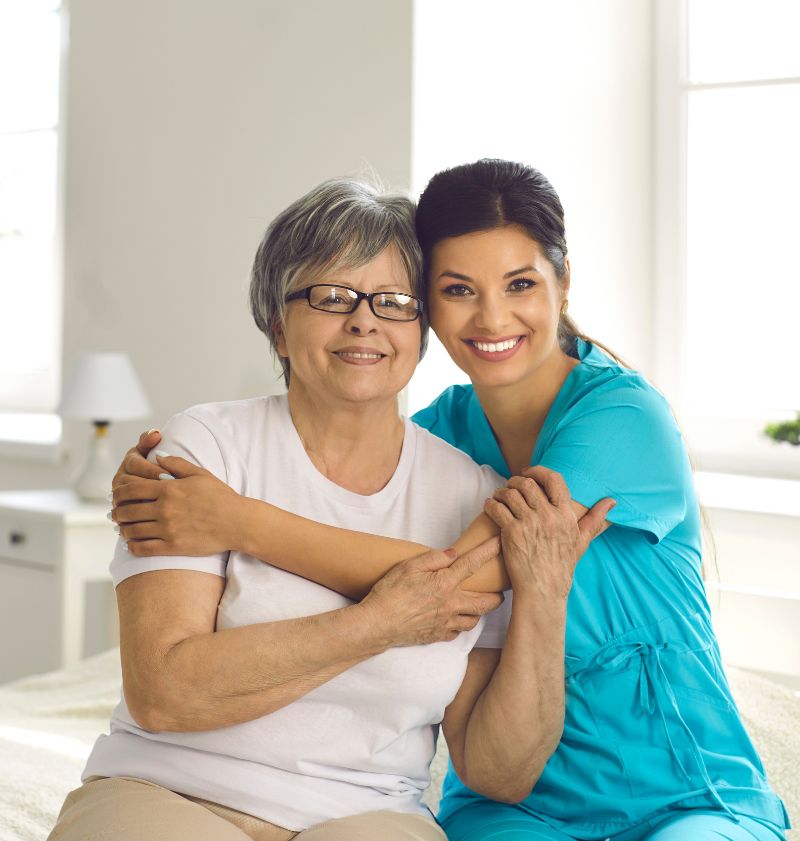 Portrait of Happy Caregiver Together with Senior Patient Sitting on Bed in Retirement Home Portrait of Happy Caregiver Together with Senior Patient Sitting on Bed in Retirement Home